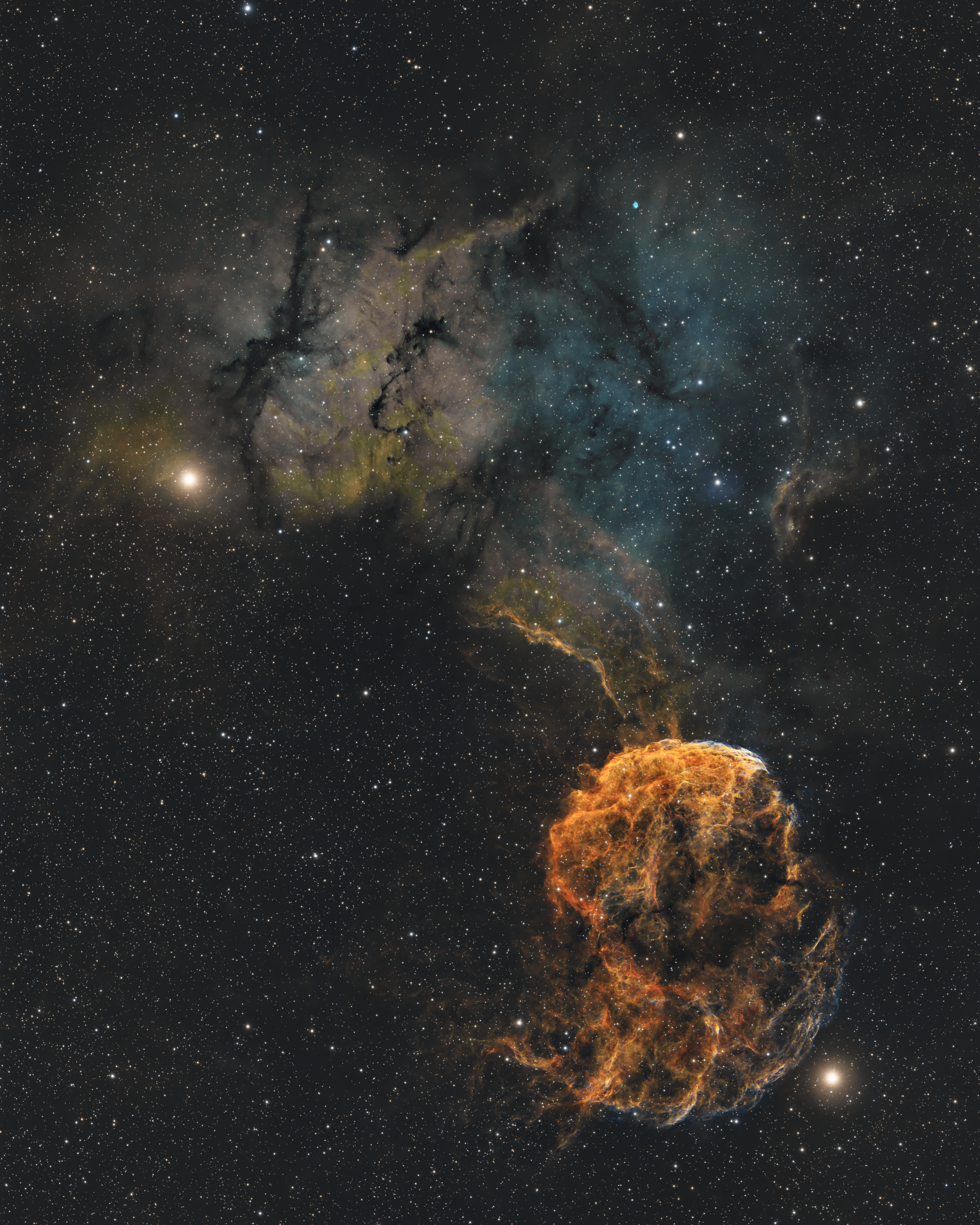 A wide starfield fills the frame, with a bright, textured orange cloud of glowing gas in the lower right that resembles a jellyfish. Its center is dense and fibrous and its lower edge frayed into wisps. Fainter strands of orange gas arc upward from it into a larger, hazy cloud that stretches across the top half of the image, fading from murky yellow-green on the upper left into soft blue on the upper right and cut through by dark, jagged dust lanes. Small white stars are scattered over the black background, with a couple of brighter yellow-white stars standing out near the left edge and below the main jellyfish-shaped cloud.