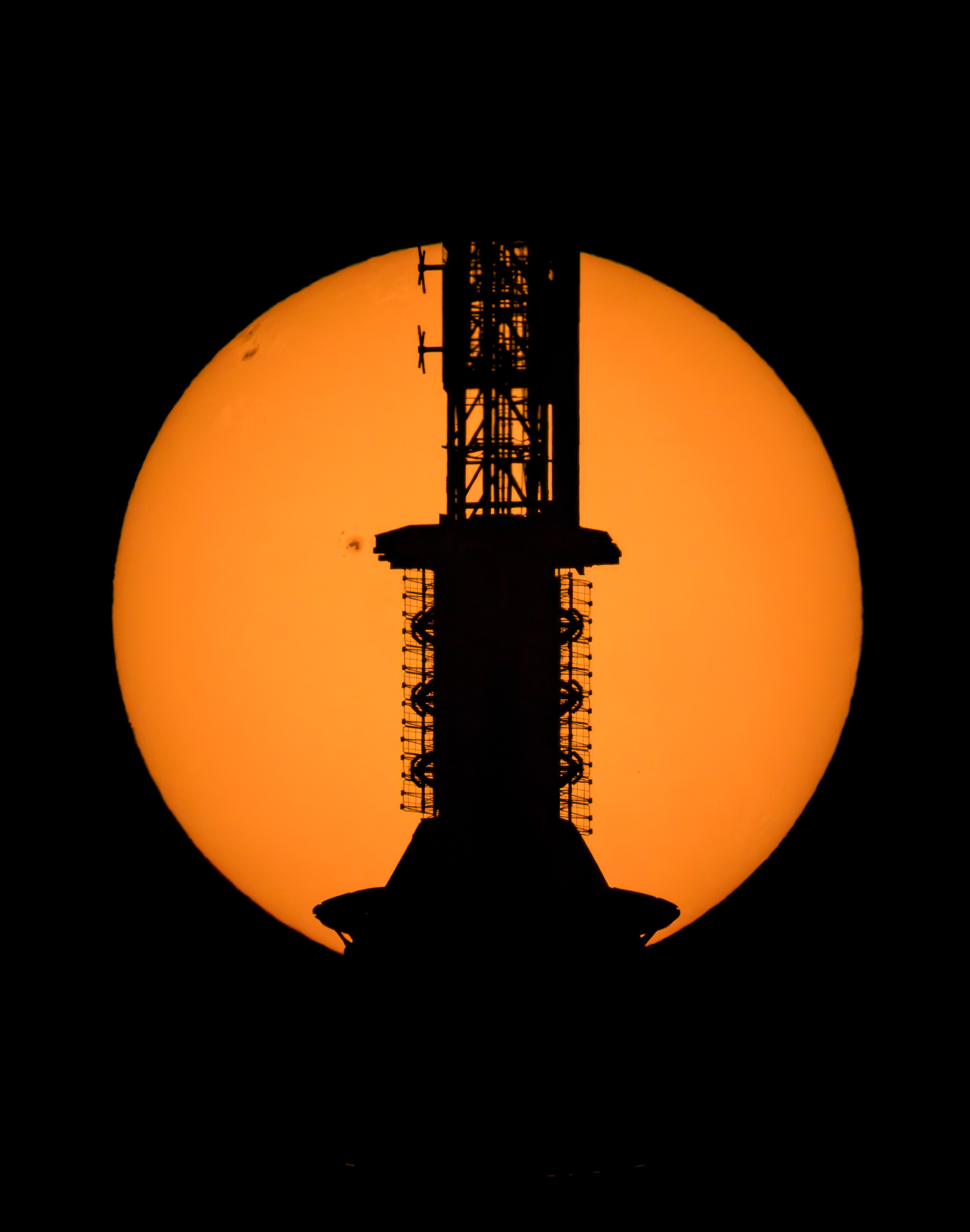 A large glowing amber disk — the Sun — fills the frame against a black sky, with a few small dark spots scattered across its left side. The dark silhouette of a tall, intricate broadcast antenna tower runs vertically down the center, crossing the face of the Sun.