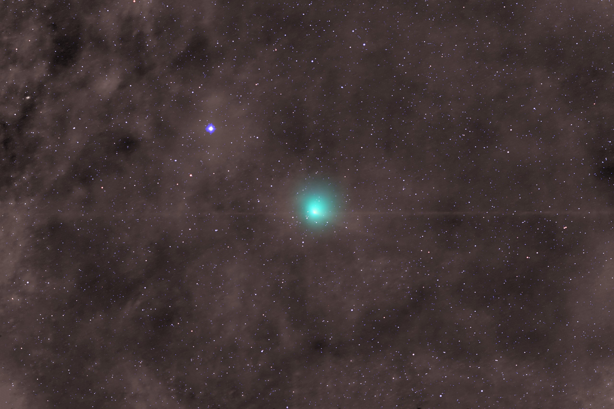 Wide-field view of deep space showing a bright teal comet near the center with a small, sharp core surrounded by a soft, round green glow. A thin, faint horizontal line crosses directly through the comet. A bright blue star with crisp four-pointed spikes shines above and to the left, set against a dense background of tiny white and reddish stars scattered over mottled brownish-grey dust clouds.