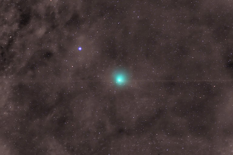 Wide-field view of deep space showing a bright teal comet near the center with a small, sharp core surrounded by a soft, round green glow. A thin, faint horizontal line crosses directly through the comet. A bright blue star with crisp four-pointed spikes shines above and to the left, set against a dense background of tiny white and reddish stars scattered over mottled brownish-grey dust clouds.
