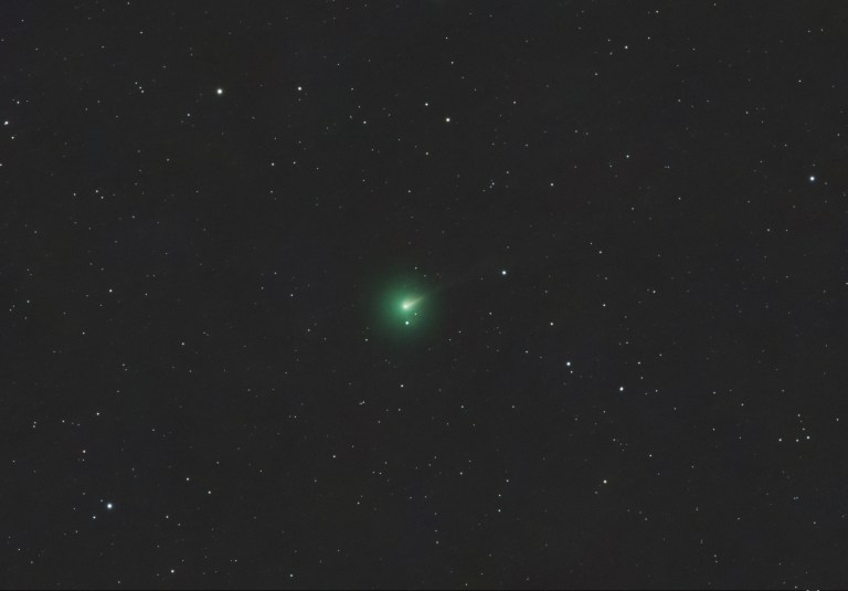A telescope image of Comet 220P in outburst, showing a bright green coma glowing near the center of the frame against a dark star-filled background. The comet's nucleus appears as a bright condensed point within the fuzzy green cloud, with a faint tail extending to the upper right.