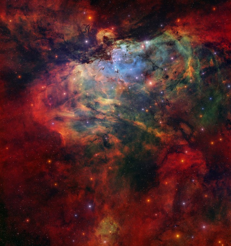 A dense, glowing nebula fills the frame, with tangled clouds of gas and dust in red, orange, green, and blue tones. Near the upper center, a bright bluish-white core and surrounding emerald and lime-green wisps stand out, while darker, finger-like dust lanes and jagged edges cut into the glowing regions. The scene is packed with stars of various sizes and colors scattered across the entire image against a darker, starry background.