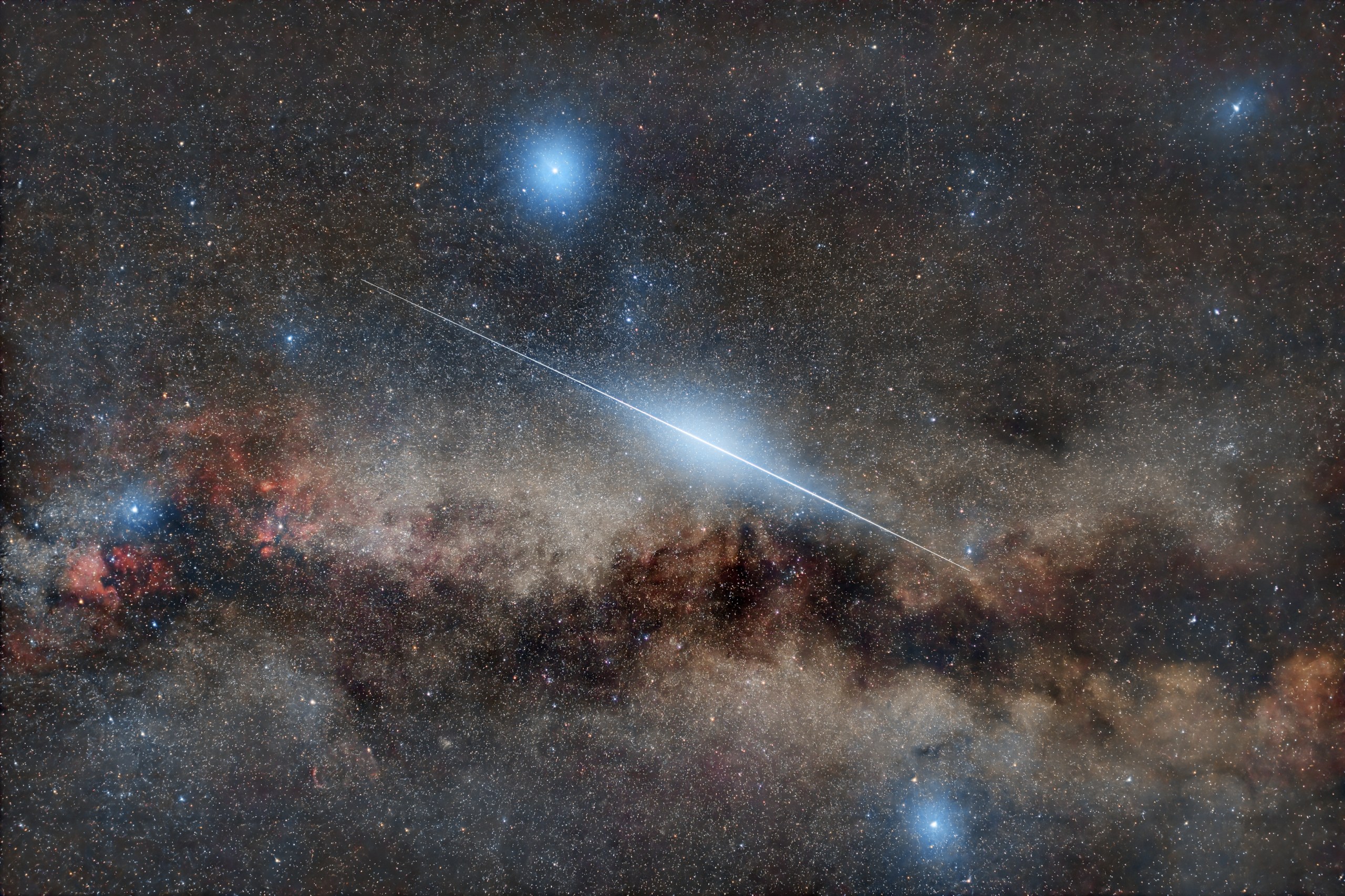 A wide, long-exposure view of the night sky shows the Milky Way stretching in a hazy, mottled band across the lower half of the frame, with creamy star clouds broken by dark dust lanes. A single bright meteor streaks diagonally from upper left to lower right as a sharp blue-white line that is brightest near its middle and fades at both ends. The background is filled with countless small stars of varying brightness, along with a few larger bluish stars that glow more strongly near the top and bottom of the image.