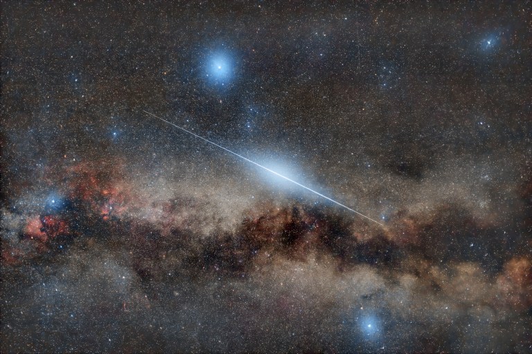 A wide, long-exposure view of the night sky shows the Milky Way stretching in a hazy, mottled band across the lower half of the frame, with creamy star clouds broken by dark dust lanes. A single bright meteor streaks diagonally from upper left to lower right as a sharp blue-white line that is brightest near its middle and fades at both ends. The background is filled with countless small stars of varying brightness, along with a few larger bluish stars that glow more strongly near the top and bottom of the image.