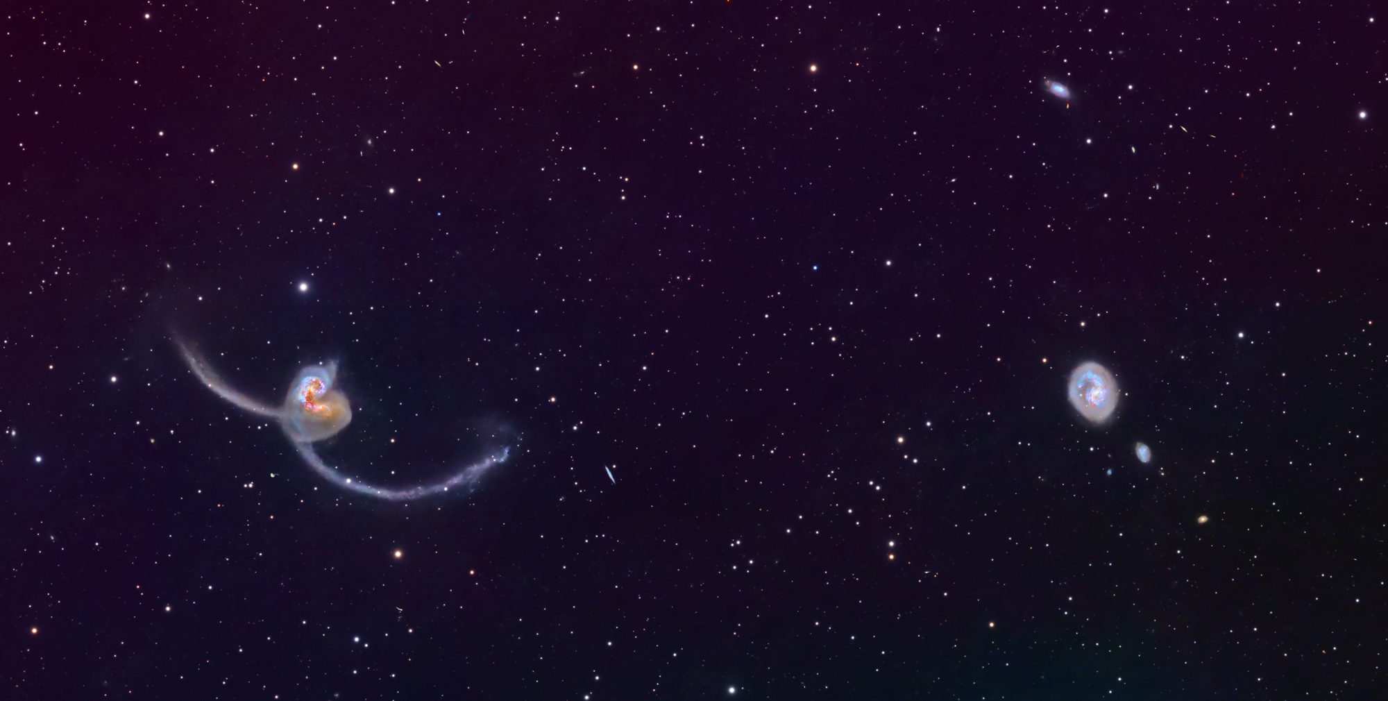 A pair of colliding galaxies sits in the lower-left of the frame, with two bright, close-together cores and long, thin arcs of bluish-white stars and gas sweeping outward in opposite directions against a dark purple-black sky. To the right, a round, face-on spiral galaxy with a bright center and faint, tightly wound arms appears above a smaller, fuzzy companion, all set in a dense background of tiny white stars scattered across the field.