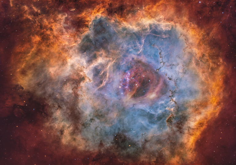 A circular, flower-like nebula fills the frame, with a bright, hollow center of soft blue and purple gas surrounded by thick, textured clouds in golden-orange and deep red. Dark, finger-like dust lanes cut into the glowing gas along the inner edges, especially on the right and bottom. A small cluster of bright stars sits near the middle of the cavity, while countless fainter stars and very subtle, reddish wisps of gas dot the darker background around the nebula.