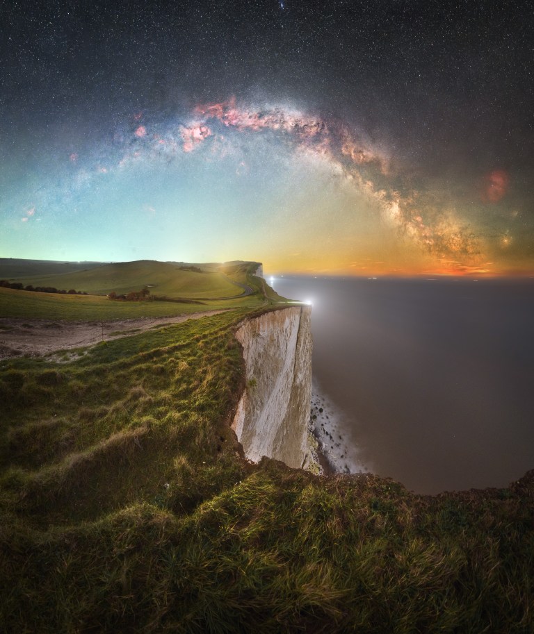 Tall white sea cliffs drop into dark, blurred water at night, with and rolling grassy hills fading into the distance on the left. Above, a dense arch of the Milky Way stretches above a colorful sky on the horizon — cool blue and teal on the left, with orange light pollution at right. The Milky Way forms bright white star clouds set against a darker sky above packed with sharp stars.