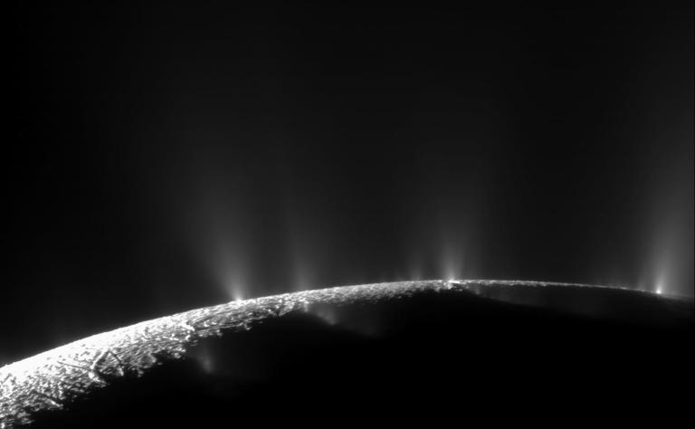 A curved, icy surface stretches across the lower portion of the frame, with multiple bright jets of vapor and particles erupting upward into a black sky from the moon's fractured southern terrain.