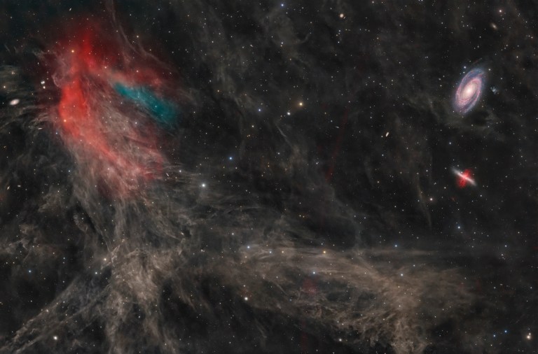 A dark starfield is veined with faint, wispy brown clouds. At left, a bright patch glows red and teal; at upper right sit a bright face-on spiral galaxy and a smaller, cigar-shaped reddish galaxy.