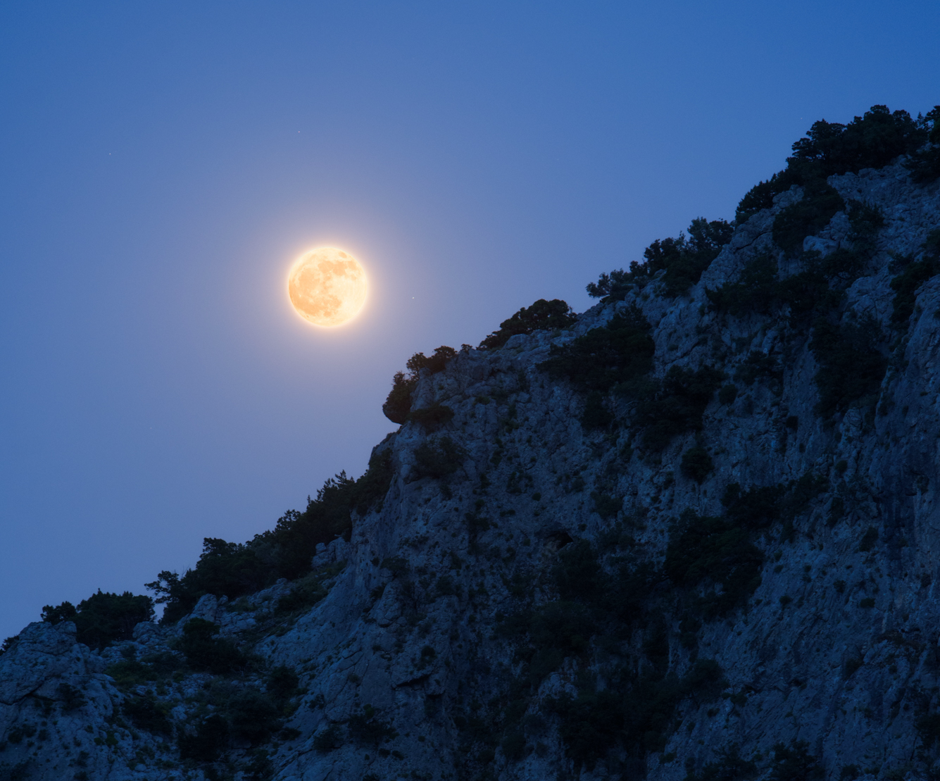 A photograph of a full Moon glowing warm orange just above the rugged ridgeline of a rocky, tree-dotted mountain slope. The Moon's surface details are visible through its amber hue, and it is surrounded by a soft golden glow against a deep blue twilight sky.