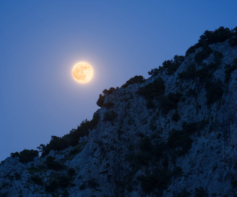 A photograph of a full Moon glowing warm orange just above the rugged ridgeline of a rocky, tree-dotted mountain slope. The Moon's surface details are visible through its amber hue, and it is surrounded by a soft golden glow against a deep blue twilight sky.