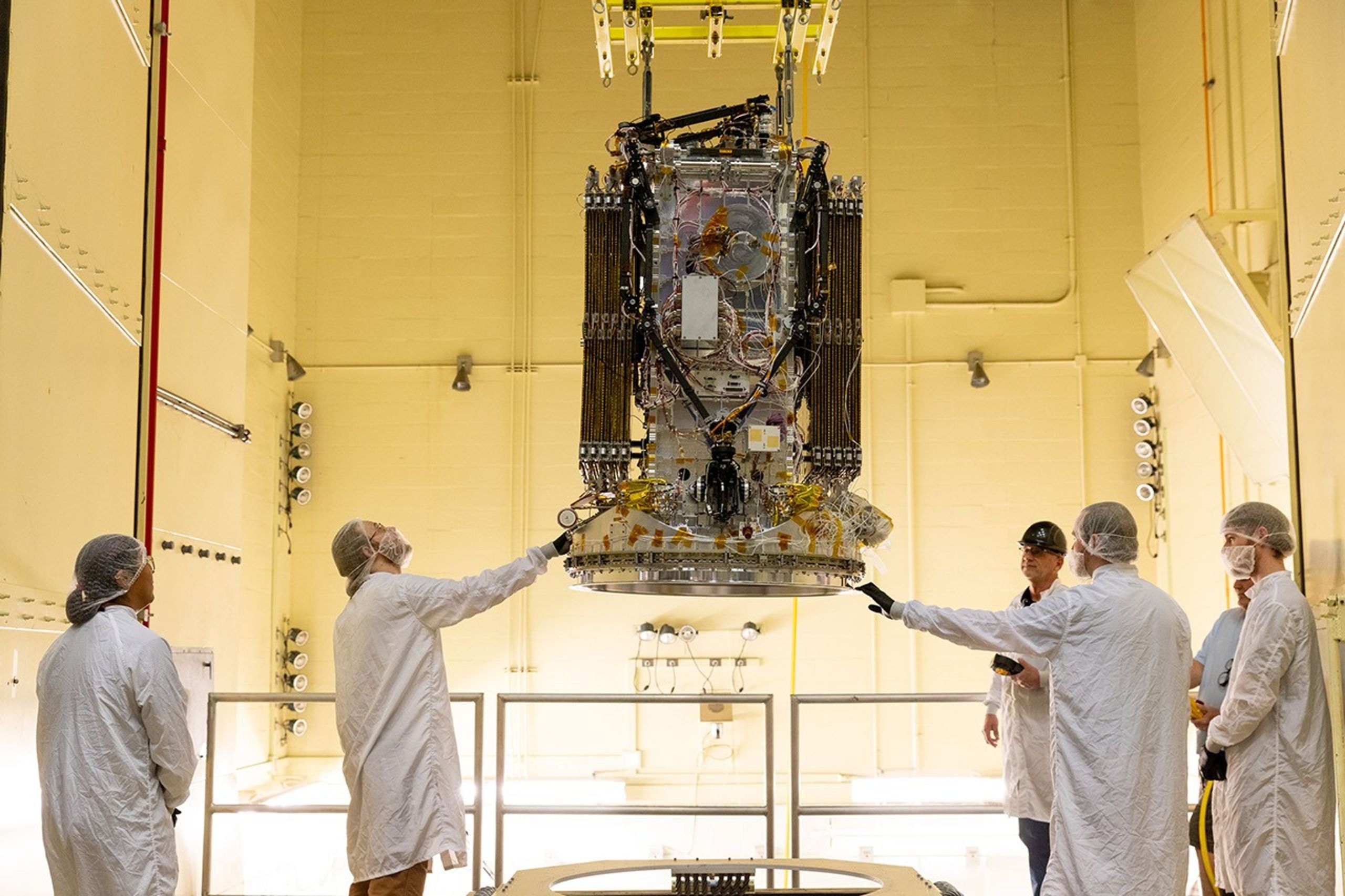 Technicians in white cleanroom suits and hairnets guide a large satellite spacecraft as it is lifted by overhead rigging inside a high-bay cleanroom facility. The spacecraft's dense exterior — packed with wiring, electronics, and gold-foil-covered components — fills the upper half of the frame, while five workers standing around a metal work stand below steady and observe it.