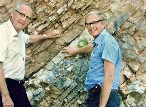 A color photograph shows two men wearing glasses standing outdoors in front of a steeply tilted, layered rock formation. The older man on the left wears a white, short-sleeved button-down shirt and dark pants, while the younger man on the right wears a blue short-sleeved shirt, dark pants, and holds a small hammer. Both men are smiling at the camera and touching the rocky, tan and grayish-brown cliff face behind them under bright daylight.