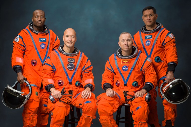 Four astronauts in bright orange Orion spacesuits pose for an official crew portrait against a muted blue-gray background. Two stand at the back and two sit in front; all hold or rest their hands near their white helmets.