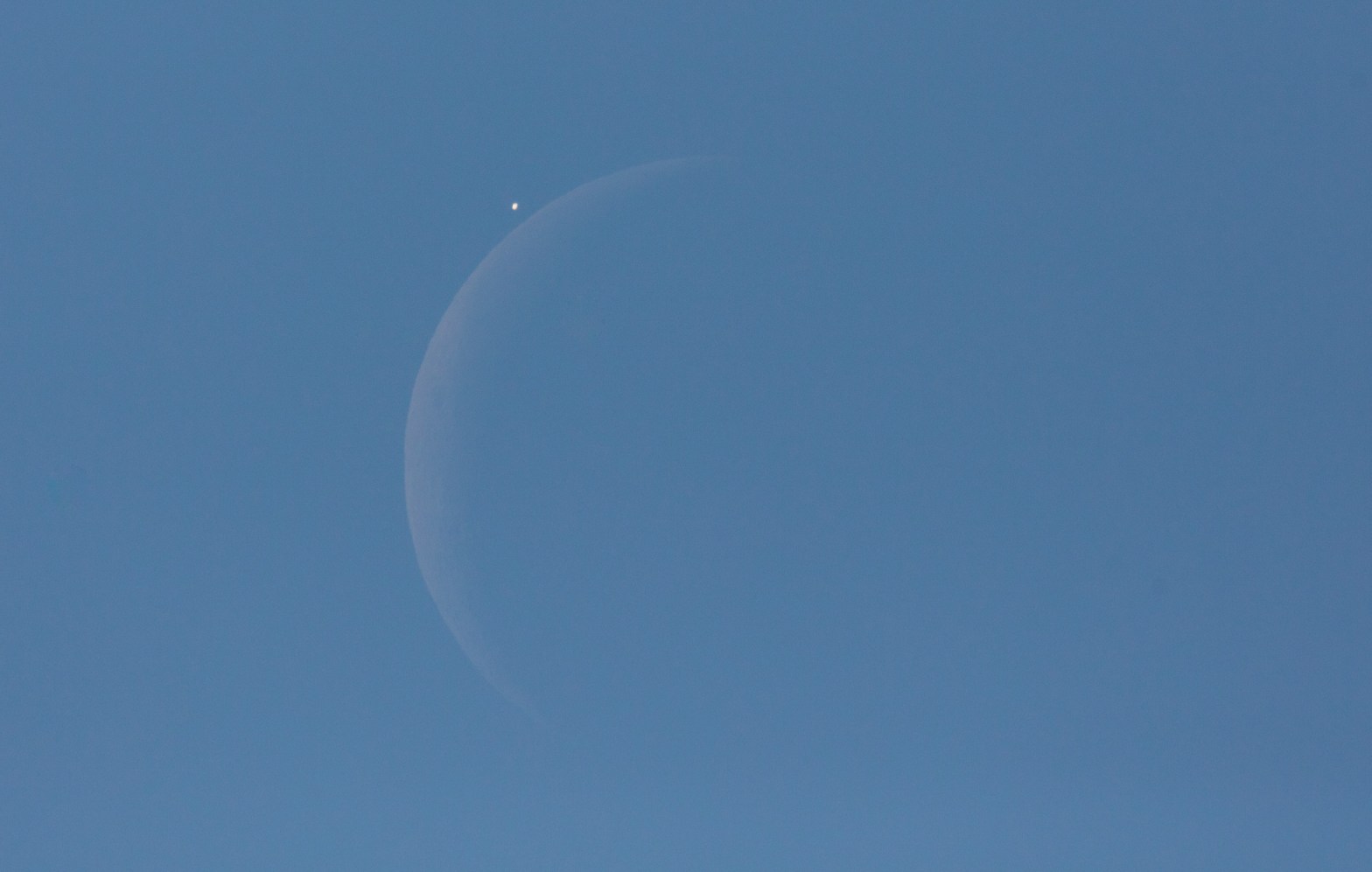 A daytime photograph of a crescent Moon visible against a clear blue sky. A bright point of light — a planet being occulted — is caught just above the Moon's illuminated limb moments before disappearing behind it, with the ghostly outline of the Moon's full disk faintly visible through earthshine.