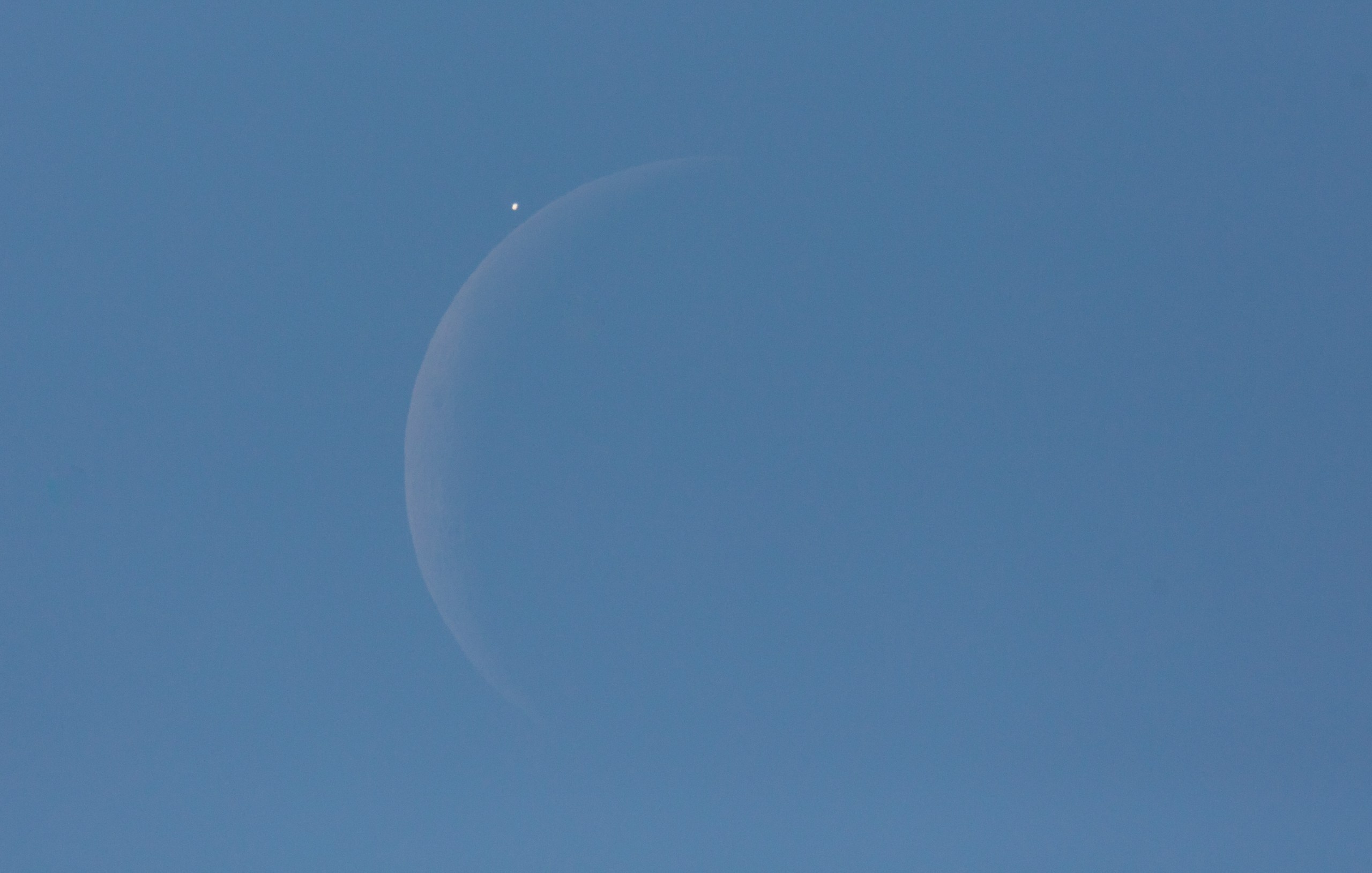 A daytime photograph of a crescent Moon visible against a clear blue sky. A bright point of light — a planet being occulted — is caught just above the Moon's illuminated limb moments before disappearing behind it, with the ghostly outline of the Moon's full disk faintly visible through earthshine.