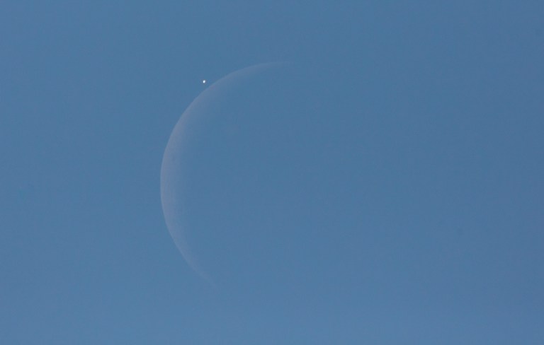 A daytime photograph of a crescent Moon visible against a clear blue sky. A bright point of light — a planet being occulted — is caught just above the Moon's illuminated limb moments before disappearing behind it, with the ghostly outline of the Moon's full disk faintly visible through earthshine.