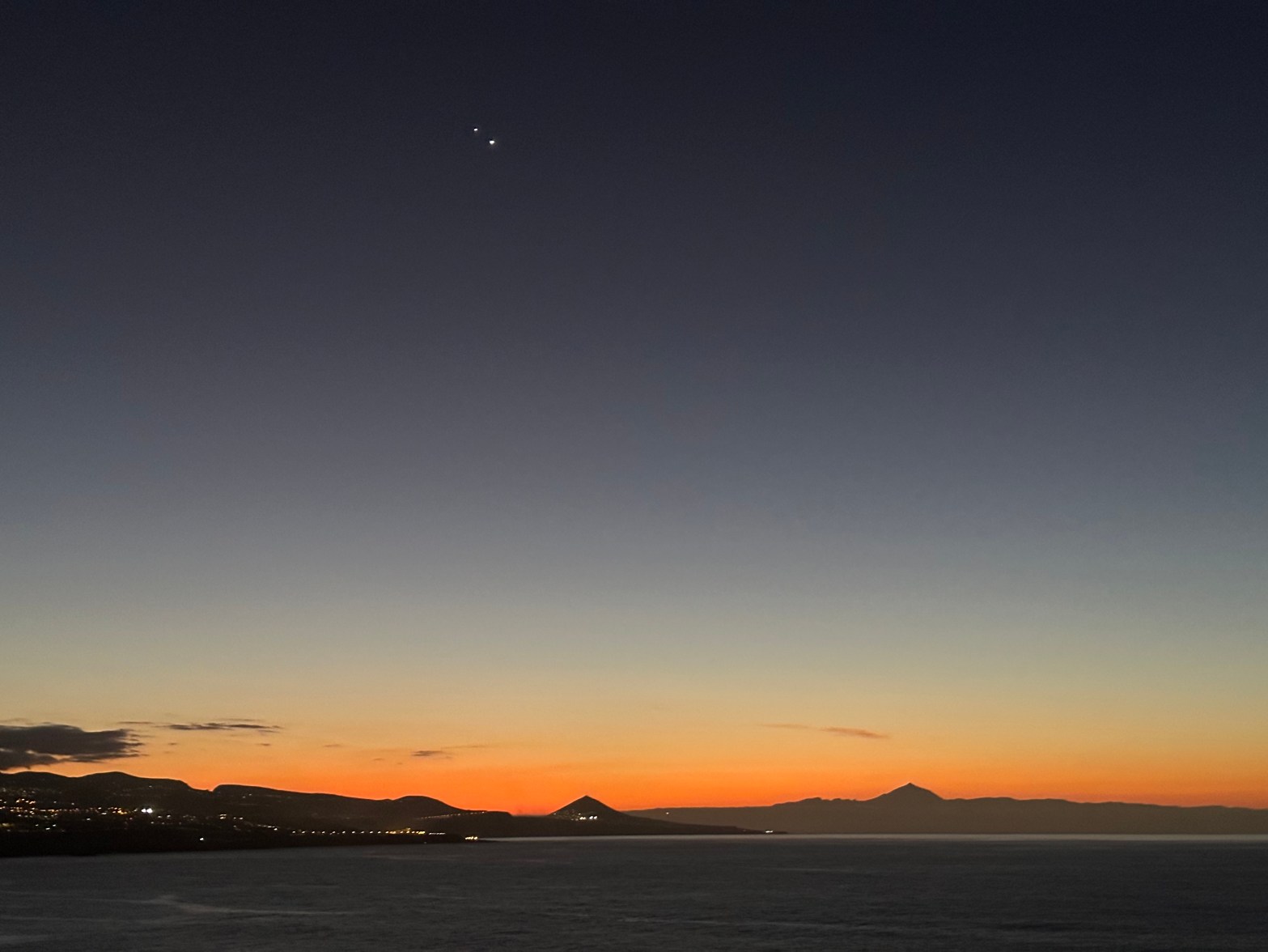 A twilight photograph taken over a calm body of water, with a vivid orange and red sunset glow along the horizon. The silhouettes of hills and a prominent volcanic peak stretch across the skyline. Jupiter and Venus appear as two bright points of light close together in the deep blue upper sky above the scene.