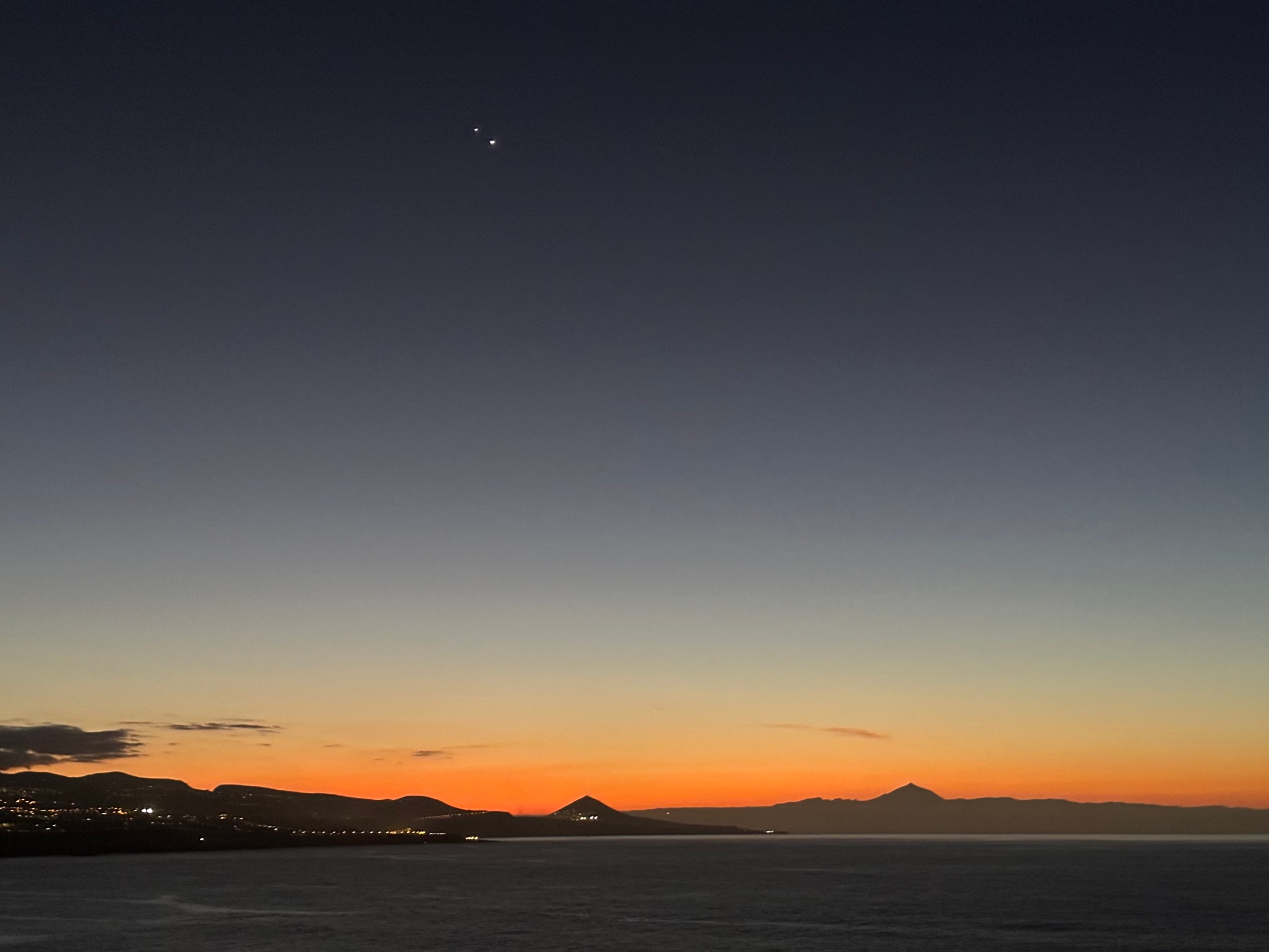 A twilight photograph taken over a calm body of water, with a vivid orange and red sunset glow along the horizon. The silhouettes of hills and a prominent volcanic peak stretch across the skyline. Jupiter and Venus appear as two bright points of light close together in the deep blue upper sky above the scene.