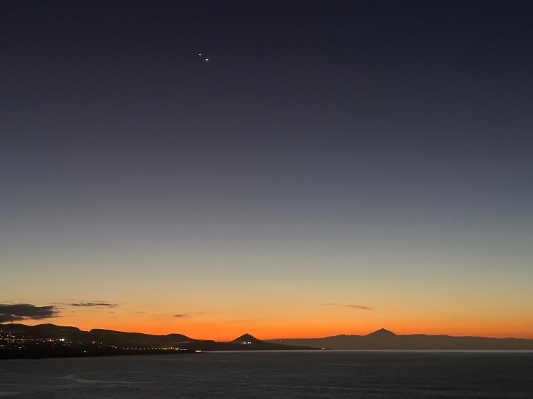 A twilight photograph taken over a calm body of water, with a vivid orange and red sunset glow along the horizon. The silhouettes of hills and a prominent volcanic peak stretch across the skyline. Jupiter and Venus appear as two bright points of light close together in the deep blue upper sky above the scene.