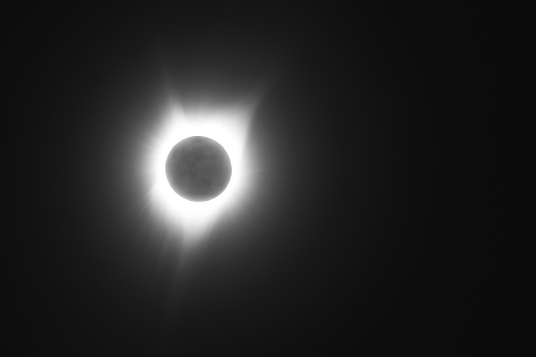 A black and white photograph of a total solar eclipse. The Moon appears as a dark circular disk at center, surrounded by the Sun's glowing white corona extending outward in streaks and rays — most prominently upward and to the lower left. The background is nearly black, emphasizing the dramatic contrast of the corona's light.