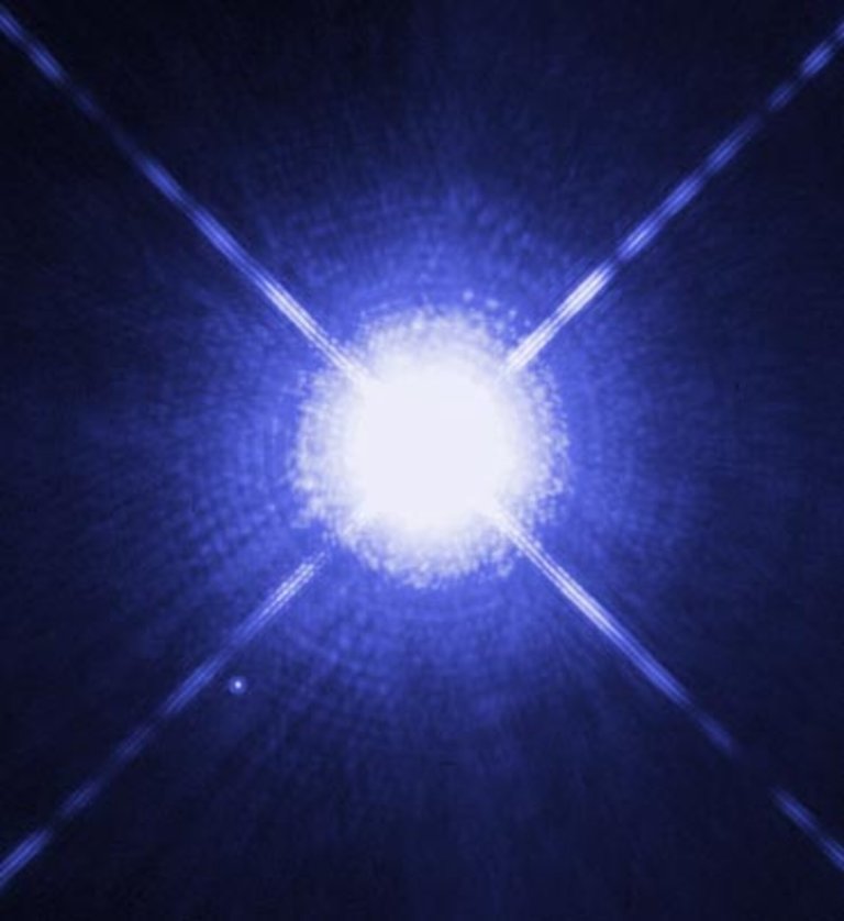 A Hubble Space Telescope image of Sirius A, the brightest star in the night sky, appearing as an intensely bright white-blue point of light with four prominent diffraction spikes extending diagonally across the dark blue background. Sirius B, its much fainter white dwarf companion, is visible as a small distinct point of light in the lower left of the frame.