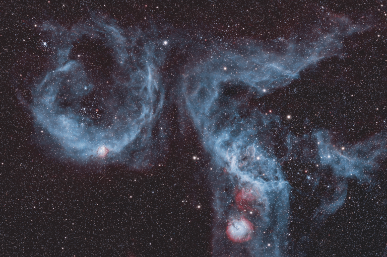 On a black, star-filled background float two large, wispy blue bubbles of gas. The rounder one at left has a small bright pink-and-white knot at its lower edge. The bigger, more tangled structure at right blends pale blue filaments with patches of red, all scattered across thousands of faint stars.