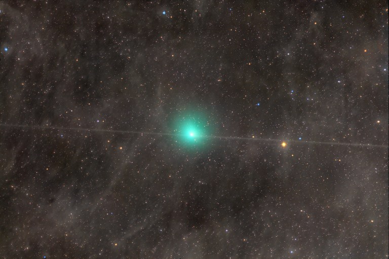 A small, bright teal-green fuzzy ball glows near the center of a dense starfield. A hair-thin straight line of light runs horizontally across the whole frame, passing through the green ball on both sides. The background holds many colorful pinpoint stars and faint brownish haze, with one bright orange star to the right.