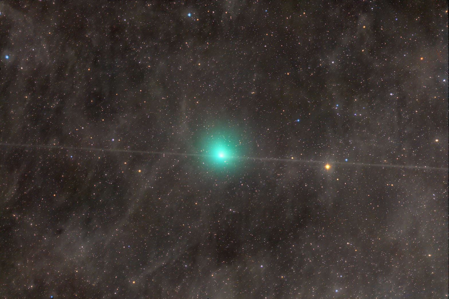A telescope image of Comet 10P/Tempel 2, showing a bright blue-green coma glowing near the center of a richly star-filled field. A diagonal satellite or aircraft trail crosses through the frame. The surrounding star field is dense with colorful blue, orange, and white stars against a dark background.