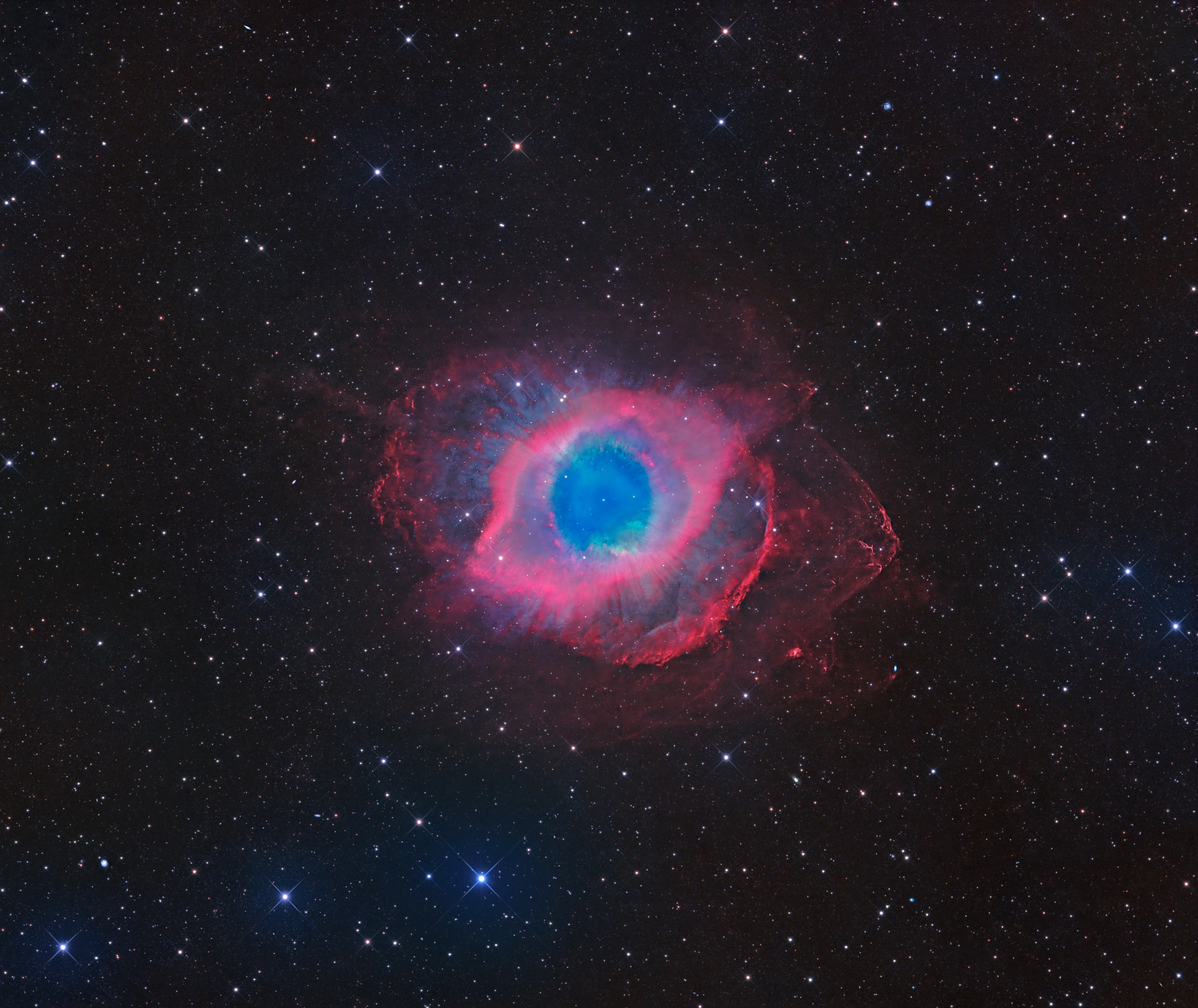 A glowing, ring-shaped cloud sits at the center of a dense star field, with a bright blue-green disk in the middle surrounded by a thick band of pink and magenta. Wispy, cloud-like red tendrils drift outward unevenly from the ring, reaching farther out toward the right side and giving the whole shape a lopsided, eye-like look. Countless small white, blue, and orange stars fill the dark background around it.