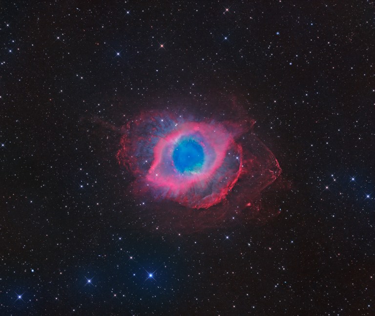 A glowing, ring-shaped cloud sits at the center of a dense star field, with a bright blue-green disk in the middle surrounded by a thick band of pink and magenta. Wispy, cloud-like red tendrils drift outward unevenly from the ring, reaching farther out toward the right side and giving the whole shape a lopsided, eye-like look. Countless small white, blue, and orange stars fill the dark background around it.