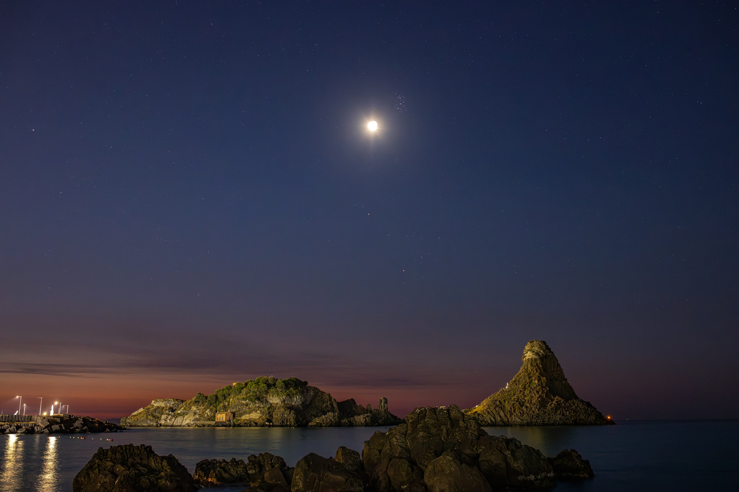 A bright crescent moon glows near the top of a deep blue night sky, with a faint cluster of stars just beside it and scattered stars across the darkness above. Below, a rocky coastline stretches across the frame — a low, tree-covered island and a tall pointed sea stack rise from calm water, silhouetted against a soft pink and orange glow along the horizon. Scattered lights dot the shoreline on the left, reflecting faintly in the still water in the foreground.