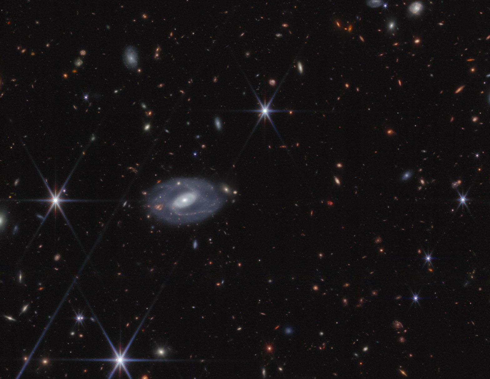 A James Webb Space Telescope deep field image showing hundreds of distant galaxies scattered across a black background in a range of shapes — spirals, ellipticals, and elongated streaks — in colors from white and blue to orange and red. A large, detailed barred spiral galaxy with a bright white core and faint blue spiral arms dominates the center-left of the frame. Several foreground stars display the distinctive six-pointed diffraction spikes characteristic of JWST imagery.