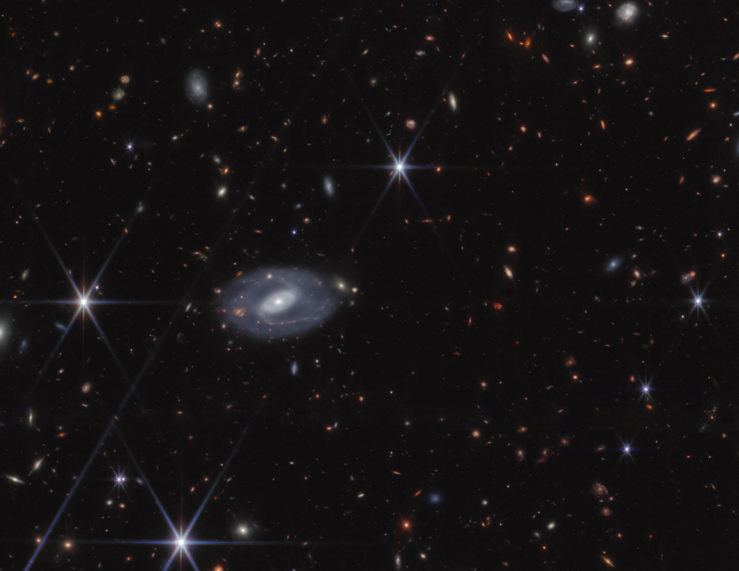 A James Webb Space Telescope deep field image showing hundreds of distant galaxies scattered across a black background in a range of shapes — spirals, ellipticals, and elongated streaks — in colors from white and blue to orange and red. A large, detailed barred spiral galaxy with a bright white core and faint blue spiral arms dominates the center-left of the frame. Several foreground stars display the distinctive six-pointed diffraction spikes characteristic of JWST imagery.