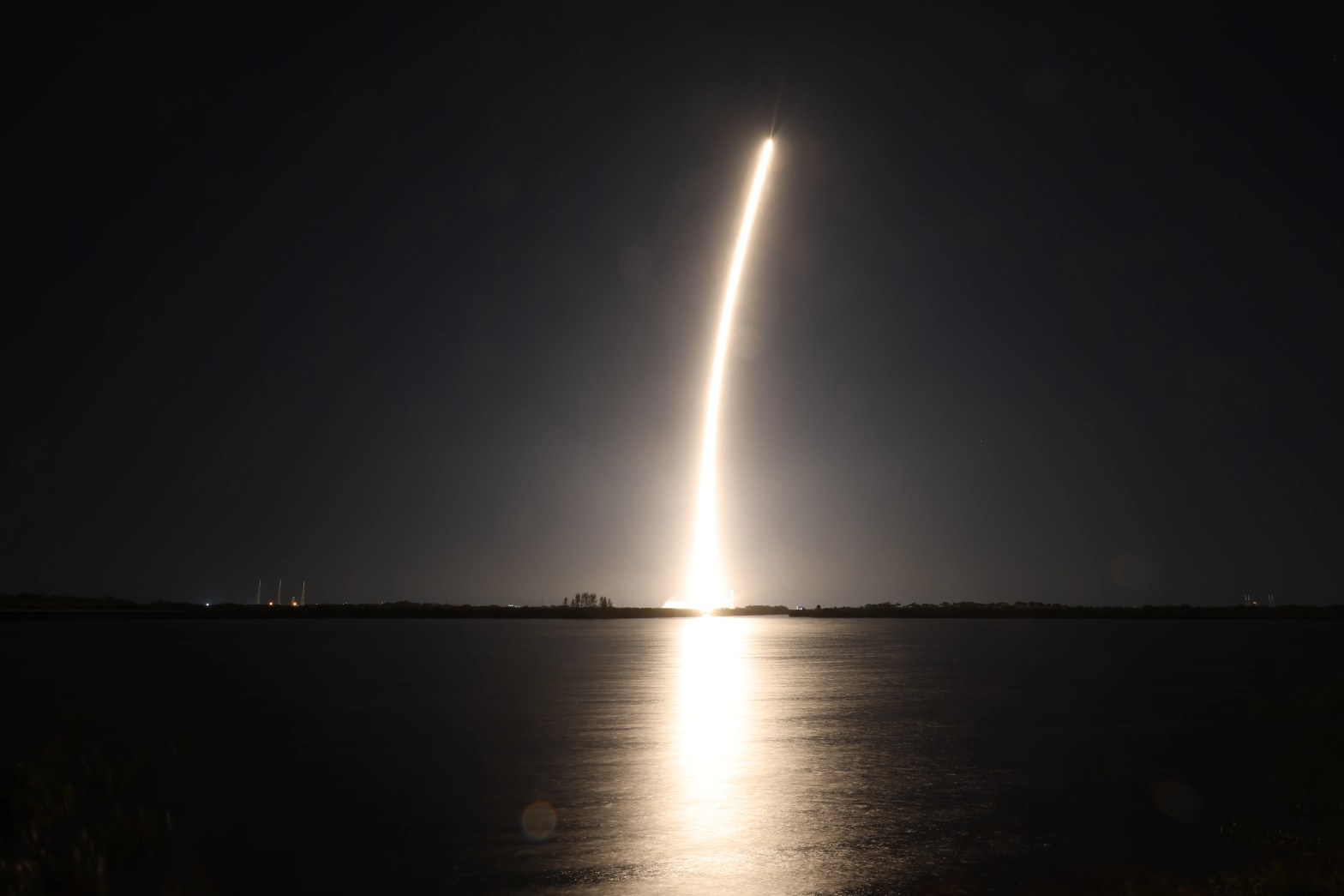 A rocket climbs into a black night sky, leaving a bright golden-white trail that curves slightly as it rises. The glow reflects off calm water in the foreground, forming a bright vertical streak on the surface, with a dark, flat horizon line separating sky and water.