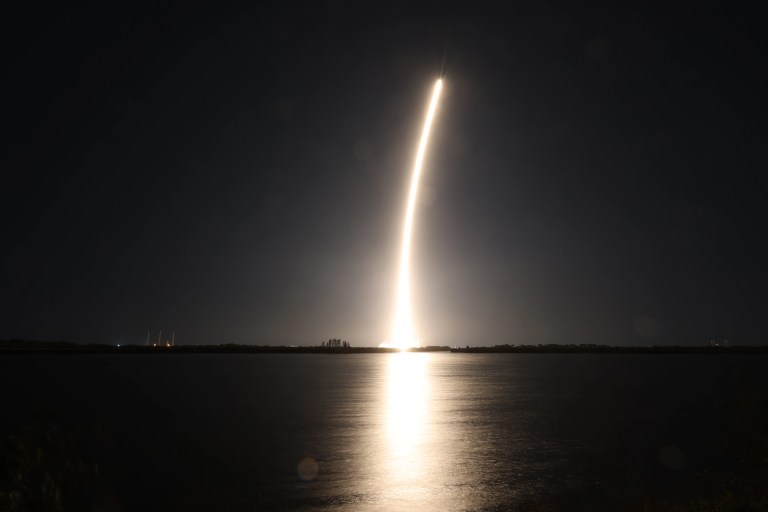 A rocket climbs into a black night sky, leaving a bright golden-white trail that curves slightly as it rises. The glow reflects off calm water in the foreground, forming a bright vertical streak on the surface, with a dark, flat horizon line separating sky and water.