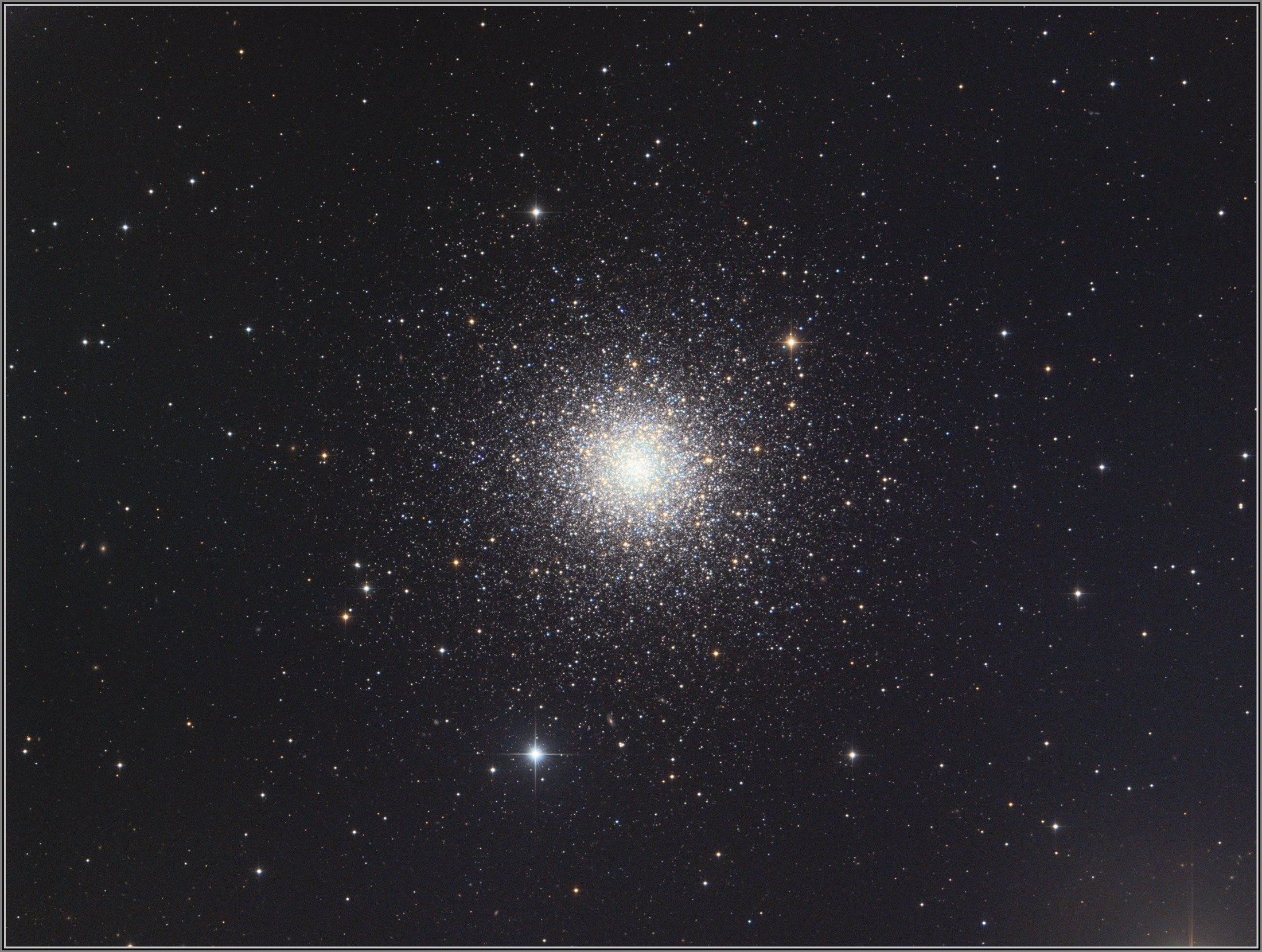 A stunning, large globular cluster (Messier 3) dominates the frame, with hundreds of thousands of stars packed into a brilliant, symmetrical sphere that glows intensely white at its core and gradually resolves into individually distinguishable stars toward the outer edges. Warm orange and cool blue-white stars are scattered throughout, becoming more densely concentrated toward the bright central nucleus. The cluster fills much of the image and is surrounded by a sparse field of foreground stars against a dark gray-black sky.