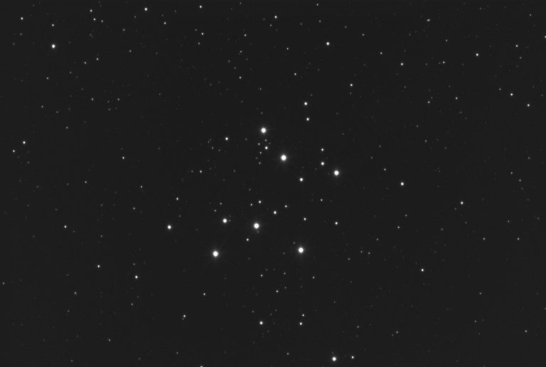 Black-and-white astrophotograph of M29, a sparse open star cluster showing roughly a dozen bright stars arranged in a loose rectangular pattern against a field of fainter background stars.