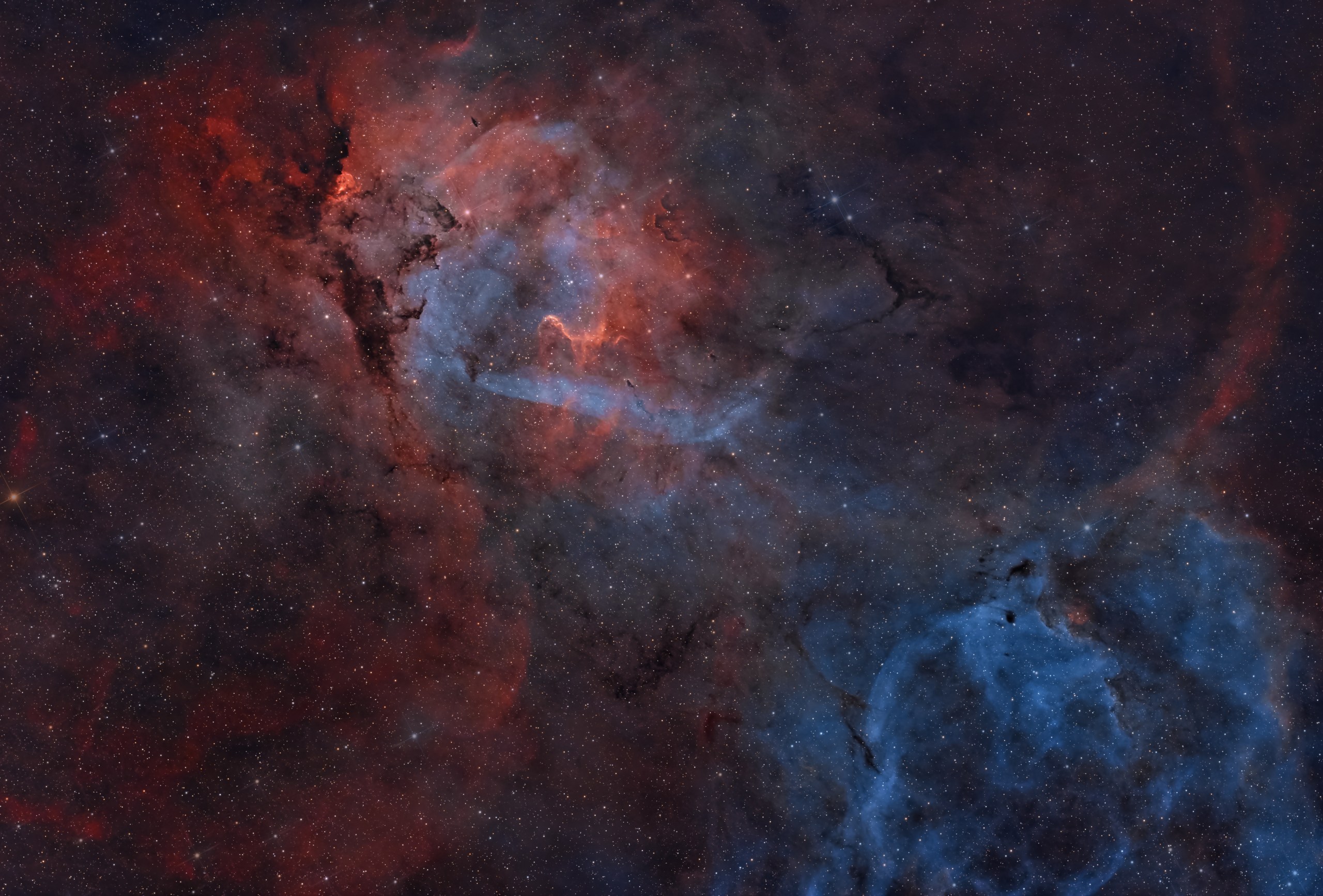 Large glowing clouds fill the frame. One deep red cloud at upper left is split by angular rays of blue light. A deeper blue cloud lies at lower right; a red arc runs up from it to the upper right corner of the frame. Together, these features appear to make up a lion’s head, body, and tail, respectively. The frame is threaded by dark, winding lanes of dust. A dense scattering of small white stars covers the whole scene.