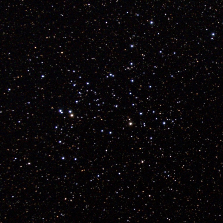 A telescopic photograph of NGC 6709, an open star cluster set against a dense, star-filled black sky. The cluster's members are scattered loosely across the frame, with a mix of bright blue-white stars and fainter orange and yellow background stars, and two noticeably brighter stars near the left-center of the cluster.