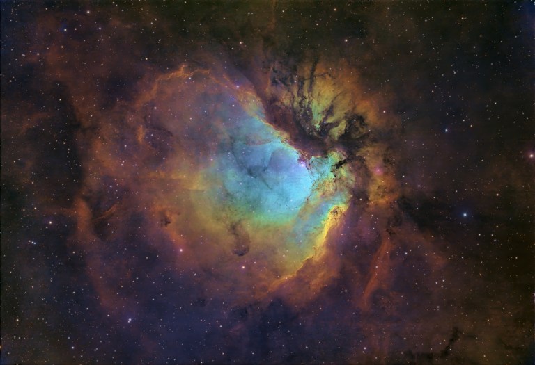 A large, glowing cloud of gas fills the frame against a dense starfield. Its center glows teal and blue, ringed and streaked by billowing gold, orange, and rust-brown clouds, with dark lanes of dust winding through the right side. The colors are vivid and painterly.