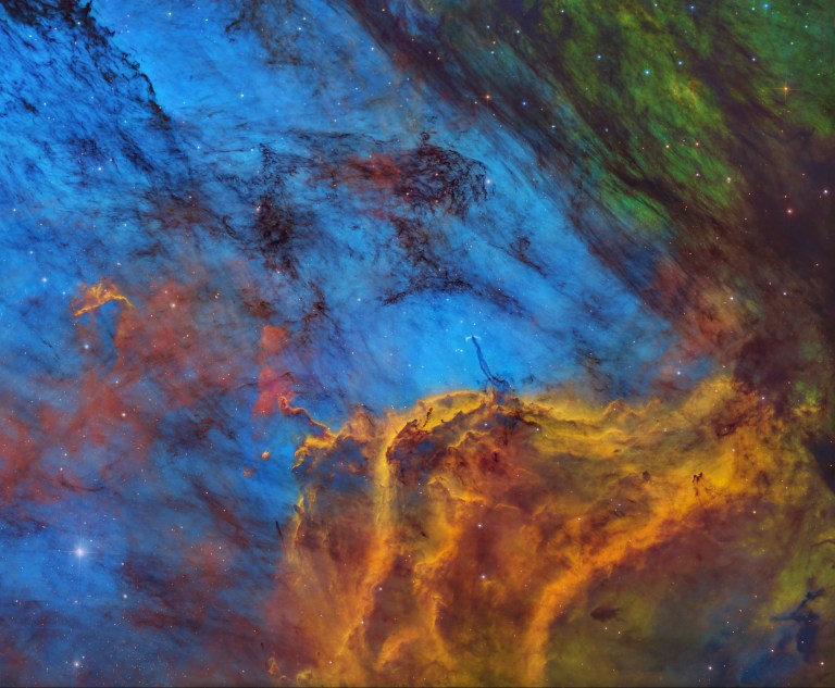 Bright orange-gold clouds billow upward across the bottom of the frame, their edges rimmed with fire against a deep blue background streaked with dark, wispy filaments. A patch of green cloud glows in the upper right corner, and a bright star shines at the lower left amid a scattering of faint stars.