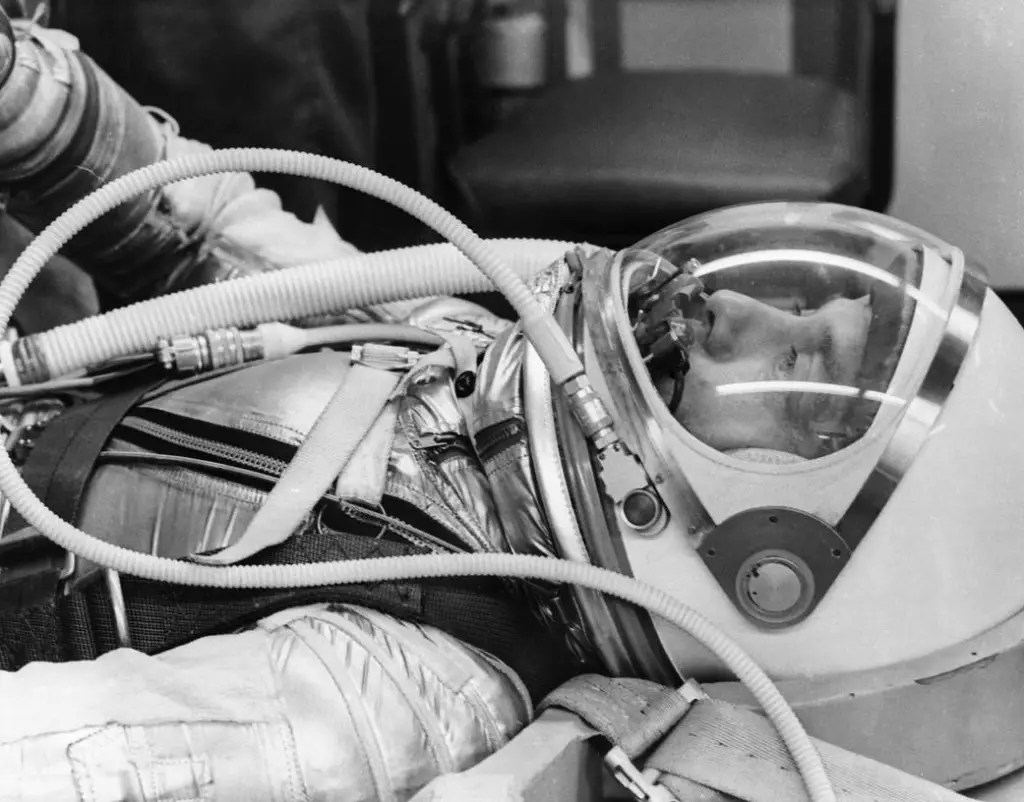 A close-up, black-and-white photograph shows a person lying flat on their back while wearing a shiny, metallic space suit and a white helmet with a clear visor. Several corrugated tubes and straps run across the torso and connect near the helmet, framing the profile of the person's face inside. The indoor setting features bright overhead lighting that reflects off the metallic suit and clear visor, with a softly blurred chair visible in the background.