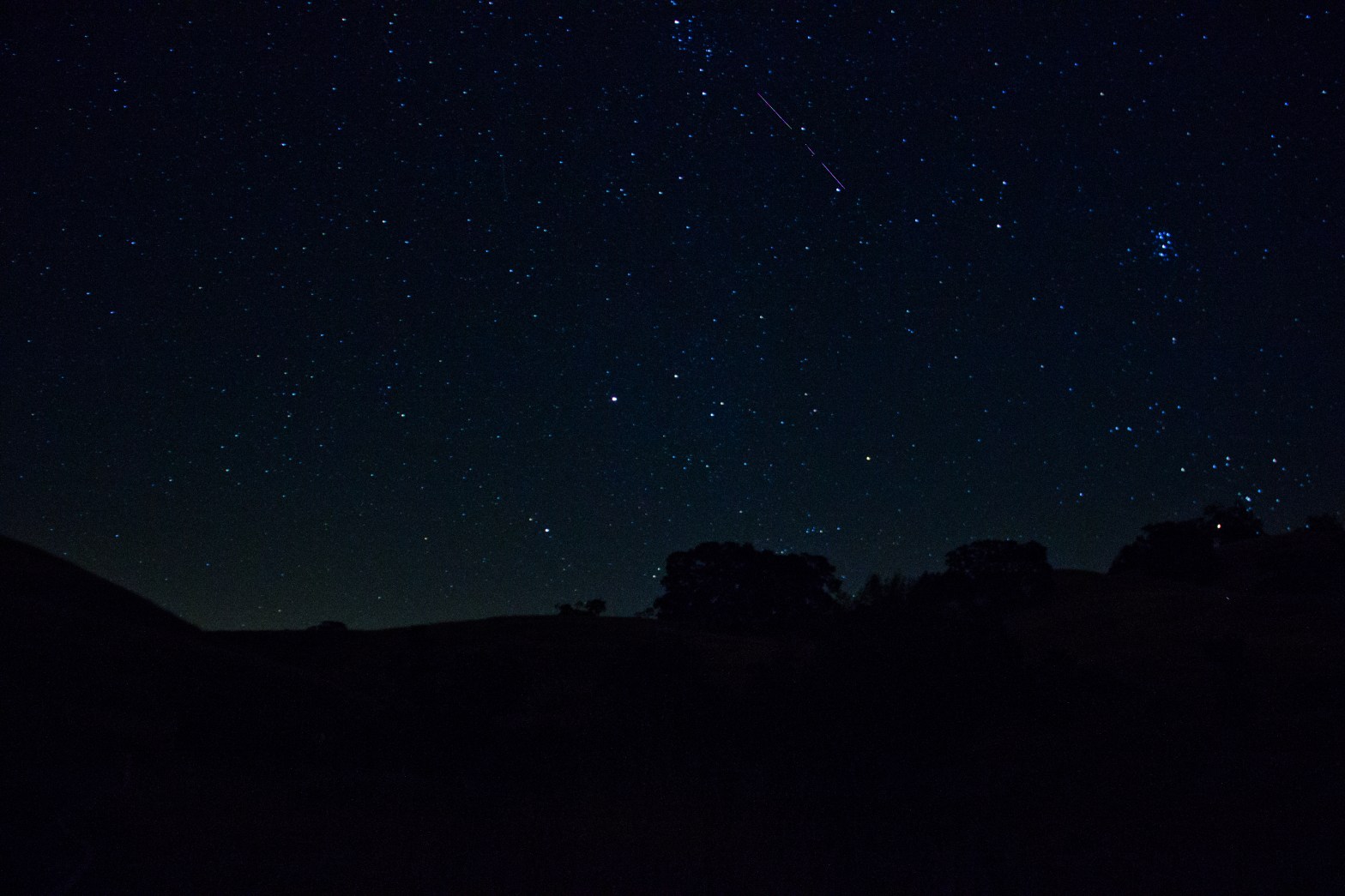 A wide-angle night sky photograph showing a lone meteor streaking diagonally across the upper portion of the frame against a deep blue, star-filled sky. The Pleiades star cluster is visible as a tight grouping of blue-white stars in the upper right. The foreground is a dark silhouette of rolling hills and trees, with a faint green glow of airglow near the horizon.