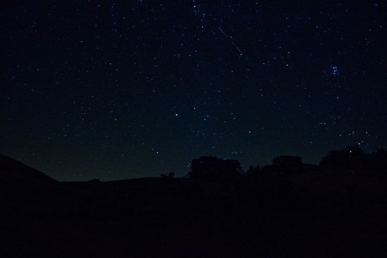 A wide-angle night sky photograph showing a lone meteor streaking diagonally across the upper portion of the frame against a deep blue, star-filled sky. The Pleiades star cluster is visible as a tight grouping of blue-white stars in the upper right. The foreground is a dark silhouette of rolling hills and trees, with a faint green glow of airglow near the horizon.