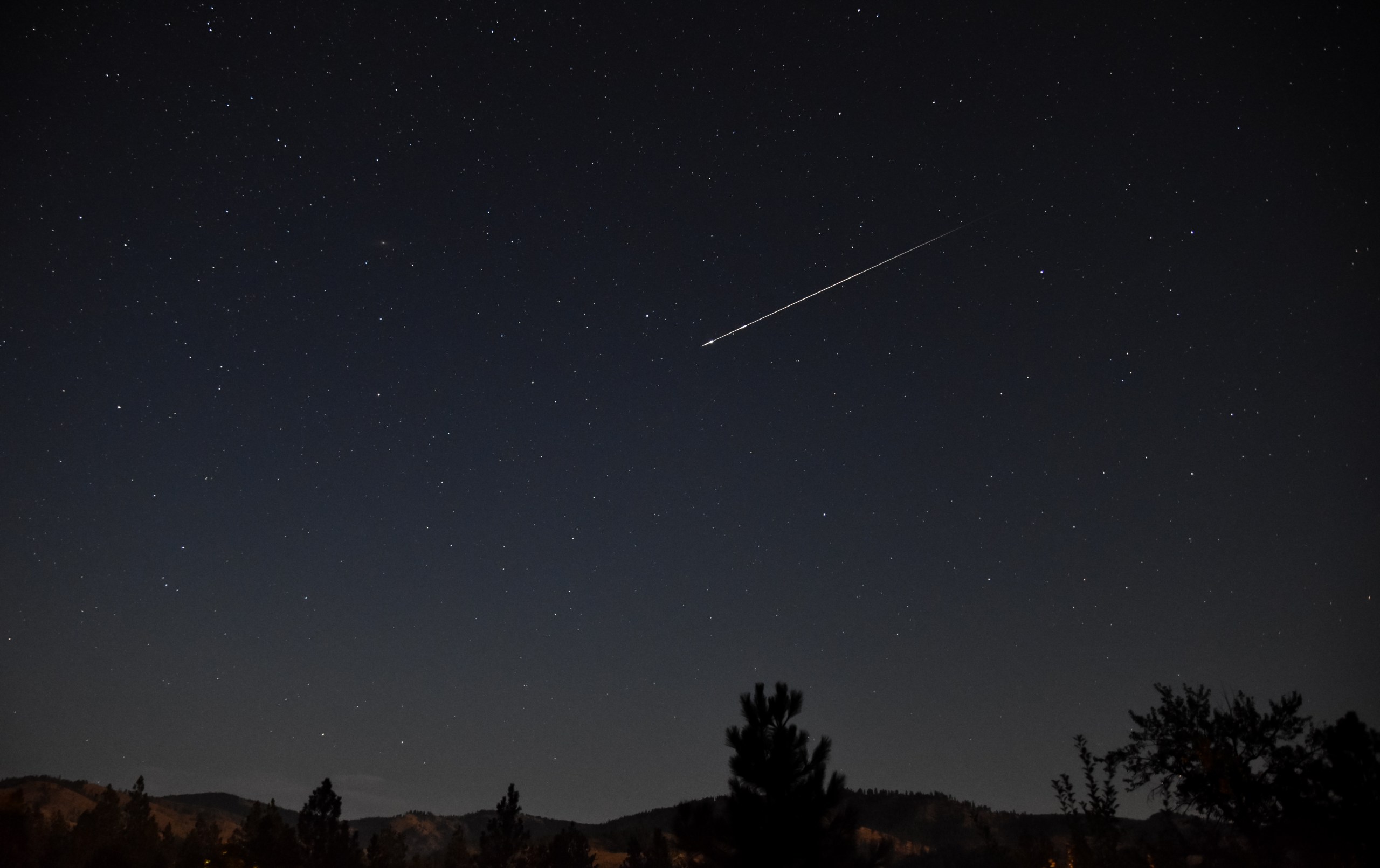 A wide-angle nightscape photograph showing a bright meteor streaking diagonally across a star-filled sky. The meteor leaves a sharp, glowing trail as it travels from upper right toward the center of the frame. The silhouettes of pine trees and rolling hills line the bottom of the image, with a faint warm glow along the horizon suggesting distant city lights or moonlight.