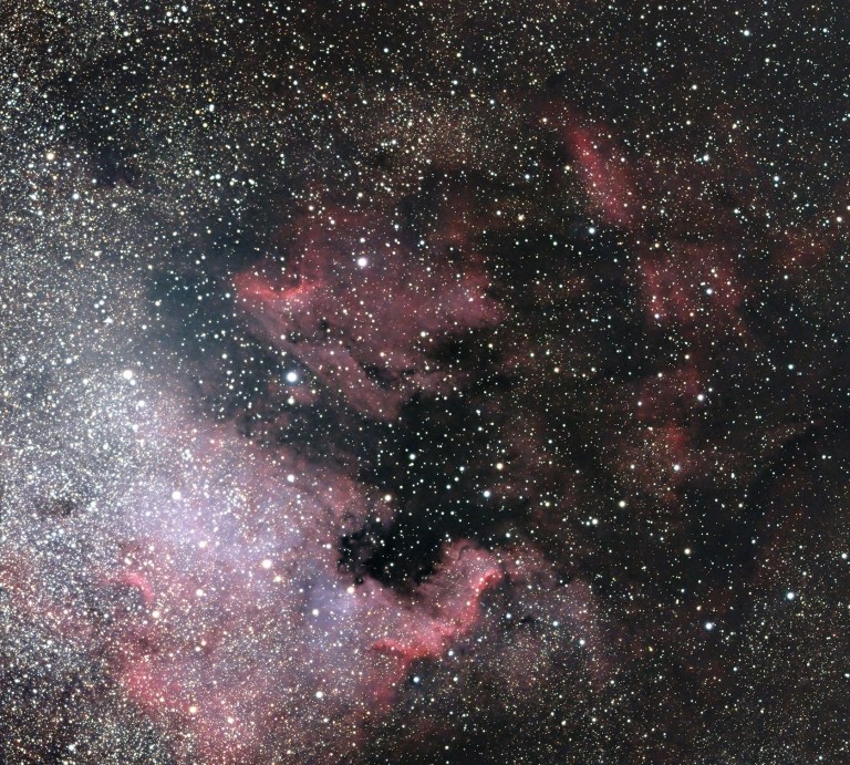 A wide-field telescopic photograph of the North America Nebula (NGC 7000) region, showing sweeping clouds of red and pink hydrogen emission gas interwoven with dark dust lanes against a densely star-filled background. A bright concentration of stars is visible along the left edge, and the characteristic shape of the nebula's dark "Gulf of Mexico" region is visible as a dark void near the center.