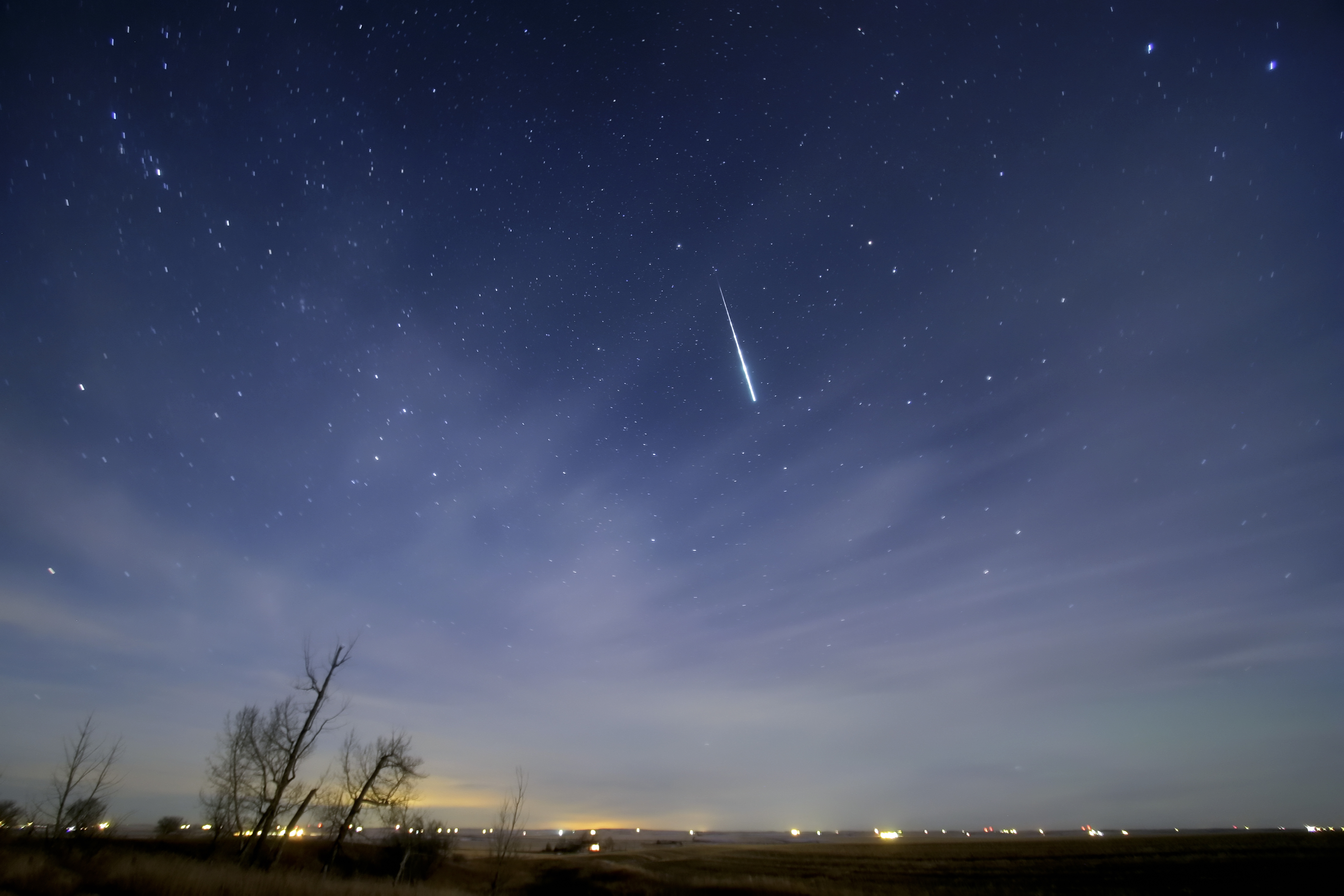 A meteor streaks down through a starry night sky over a dark prairie landscape lined with bare trees and distant scattered town lights.