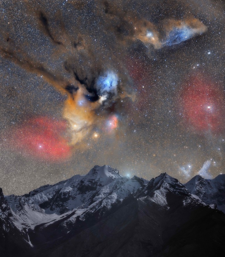 A jagged, snow-covered mountain range sits beneath a dense, star-filled sky. Above the peaks, patches of orange, red, and blue glow softly amid dark, wispy clouds of dust, with a dense cluster of yellow stars and a bright orange-white star shining just above the highest summit.
