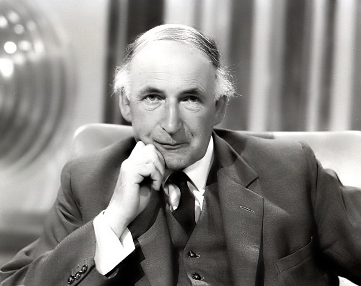 A black-and-white portrait features a man in a three-piece suit and tie seated in the center frame, resting his chin on his right hand while looking directly forward. Bright studio lighting illuminates his face and light-colored shirt cuffs, creating smooth contrast against his dark jacket and vest. The background consists of soft-focus, vertically striped curtains and a blurred metallic sphere to the left.